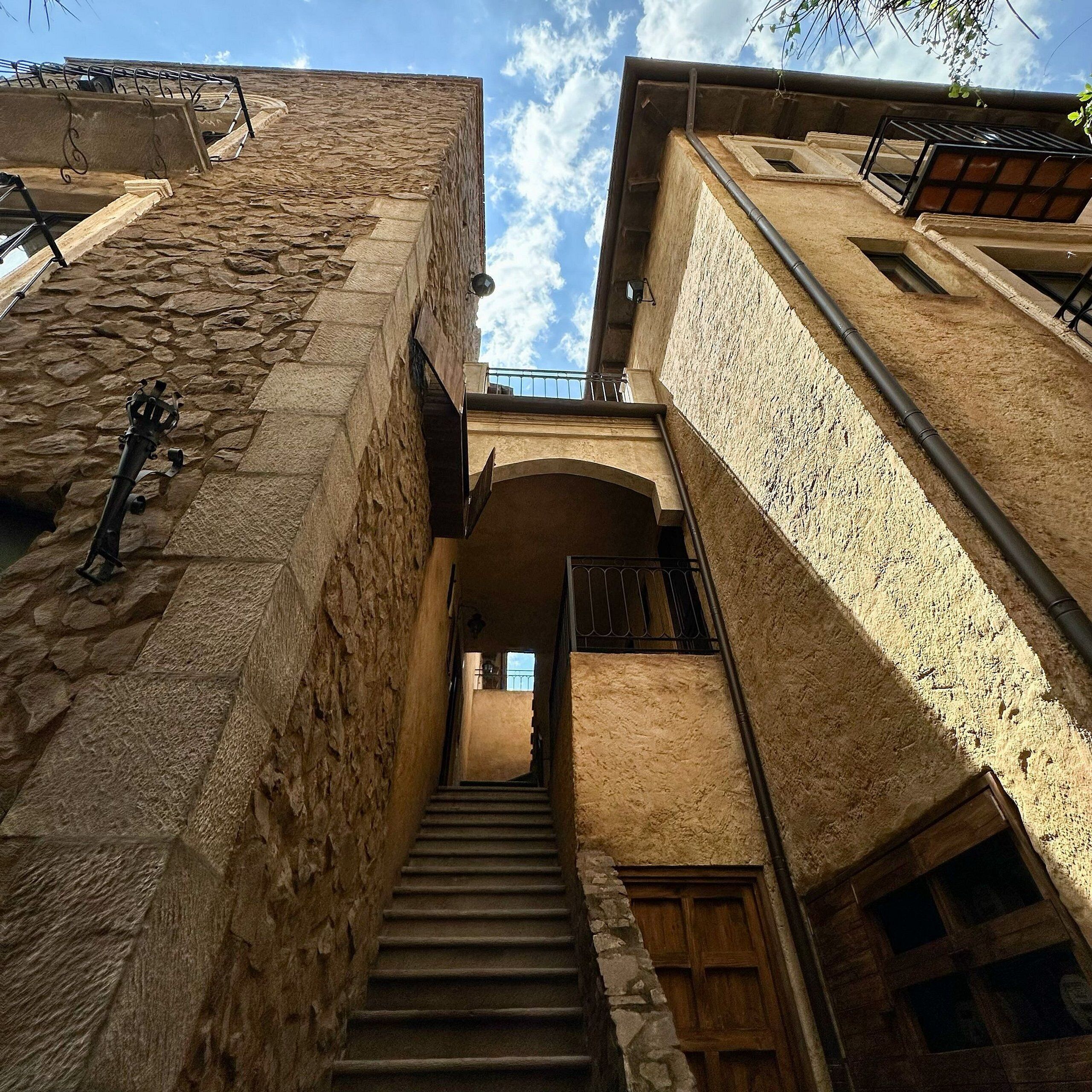 Uma escadaria de pedra estreita leva a um andar superior, ladeada por edifícios com varandas e um céu claro acima.
