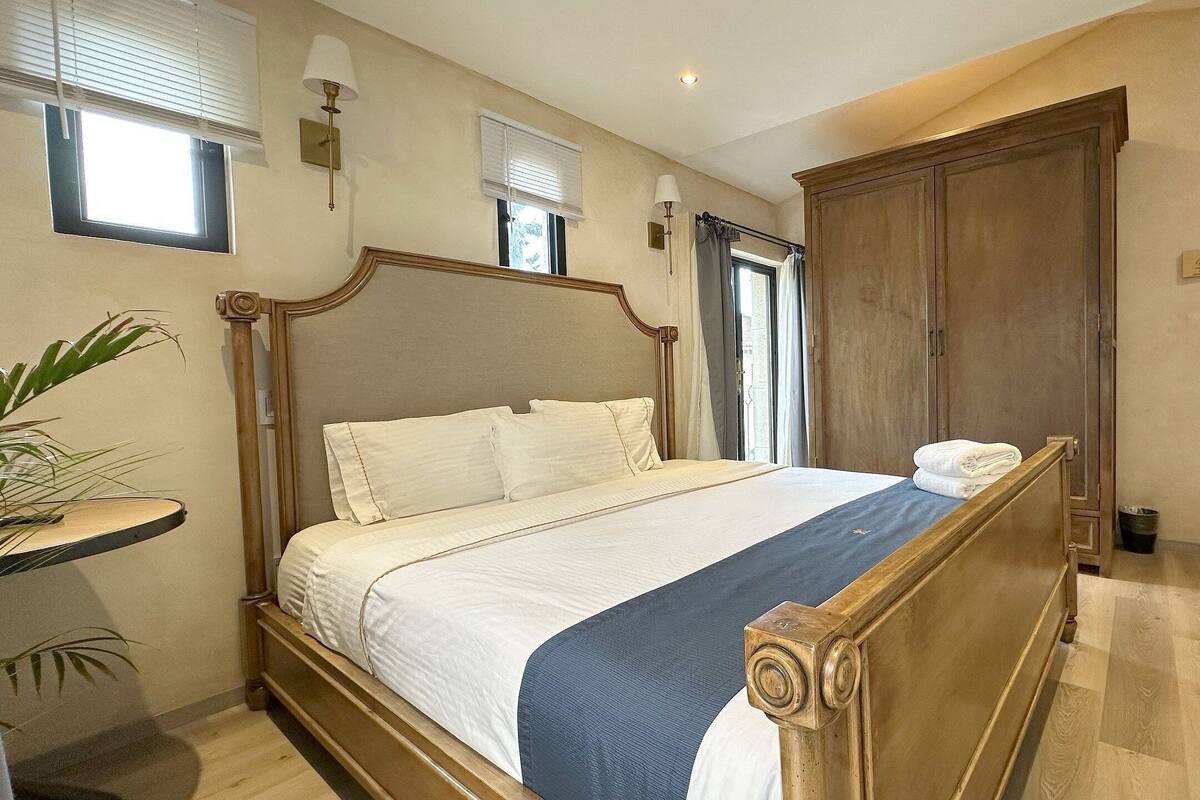 A bedroom with a large bed, a wooden wardrobe, and a small bedside table with a lamp.