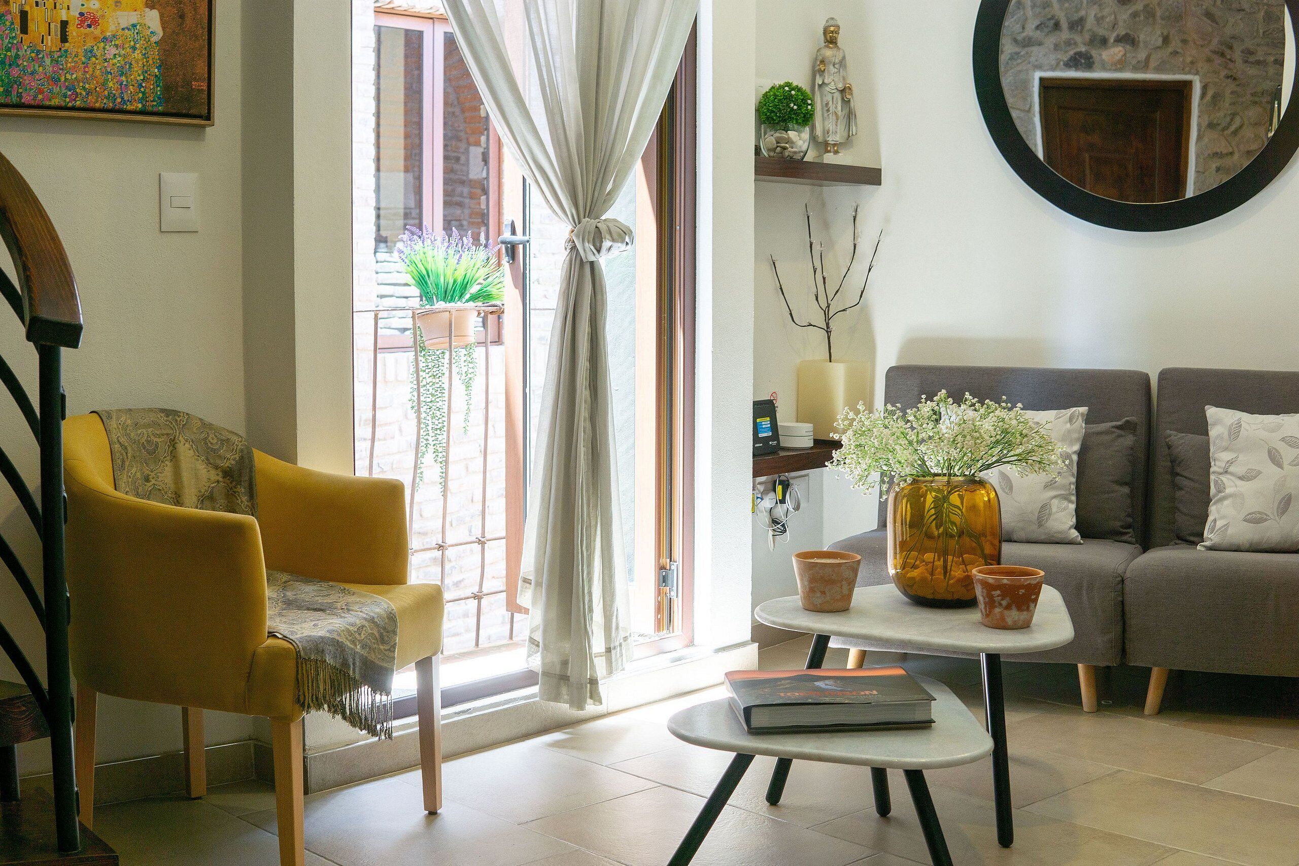 Een woonkamer met een gele fauteuil, een grijze bank, een salontafel met een boek en een vaas met bloemen.