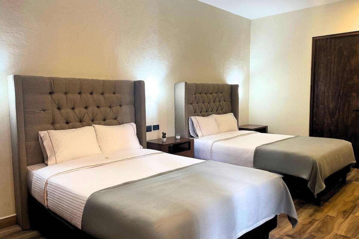 A hotel room with two beds, each with a tufted headboard, a small bedside table with a lamp, and a dark wooden floor.