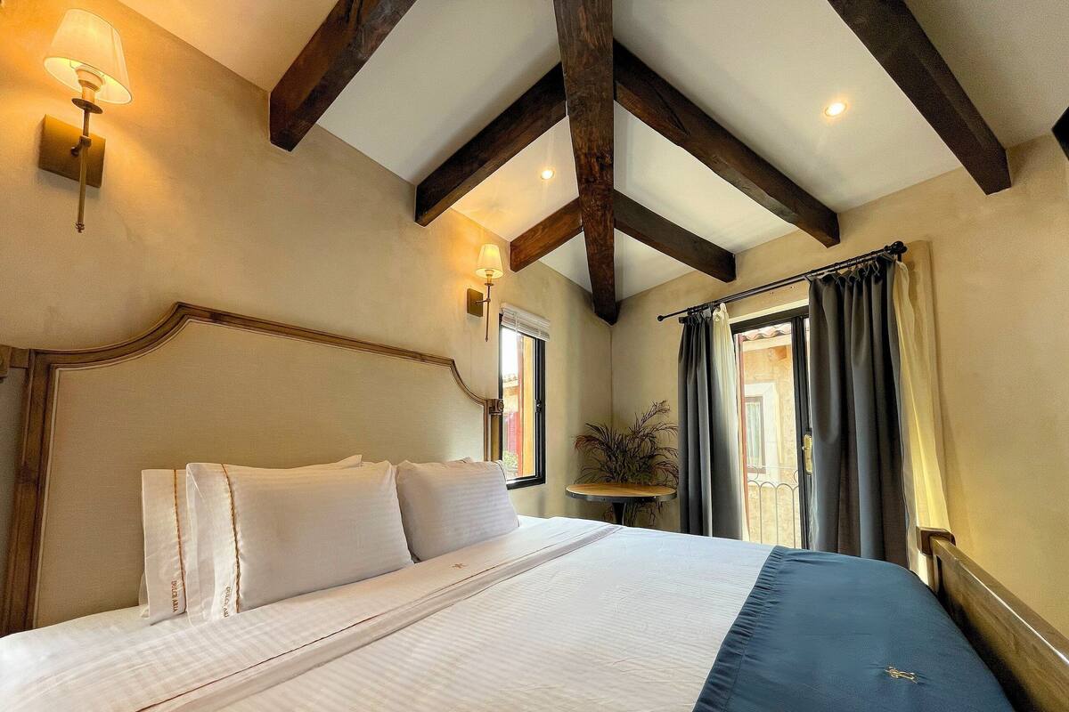 A bedroom with a large bed, wooden beams, a window with curtains, and a nightstand with a plant.