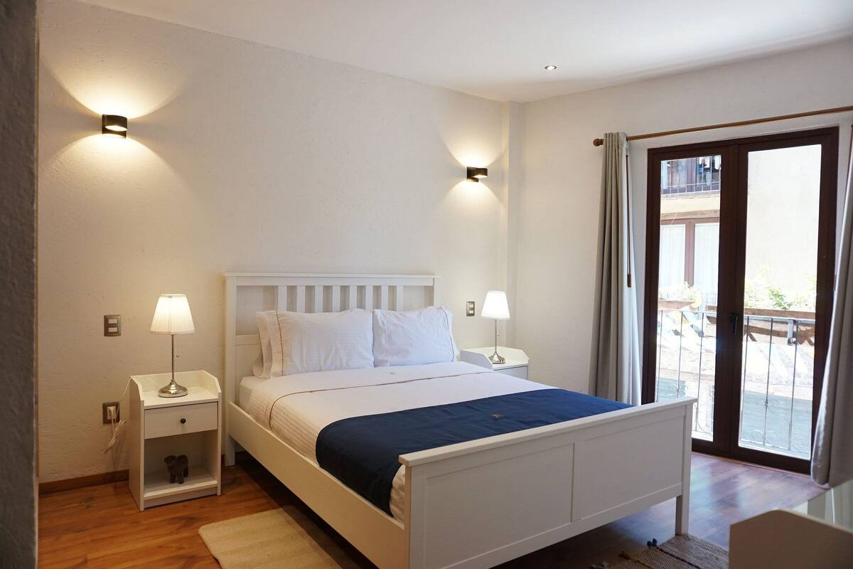 A bedroom with a bed, two bedside tables with lamps, a nightstand, a window with curtains, and a view of the outside.