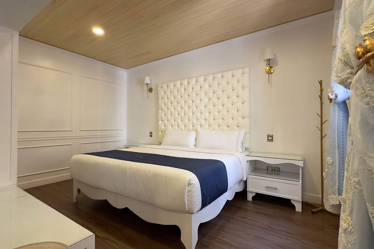 A bedroom with a tufted headboard, a bed with white and blue bedding, a nightstand with a lamp, and a decorative wall panel.