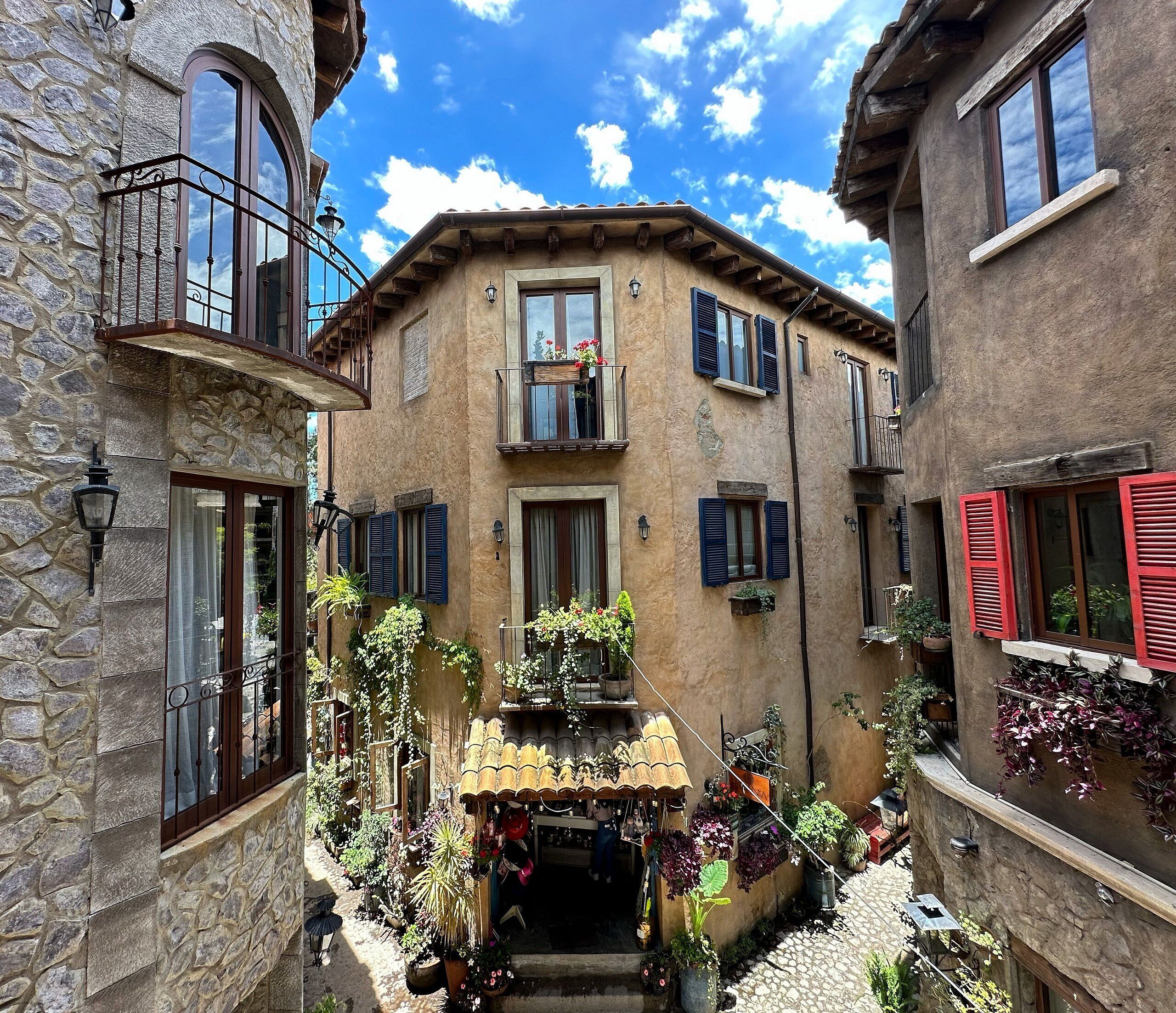 A narrow street with stone buildings, balconies, and potted plants.