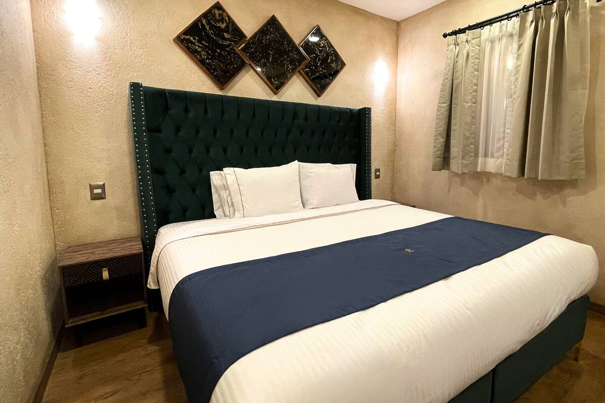 A neatly made bed with a dark headboard, a bedside table, and a wall-mounted light fixture.