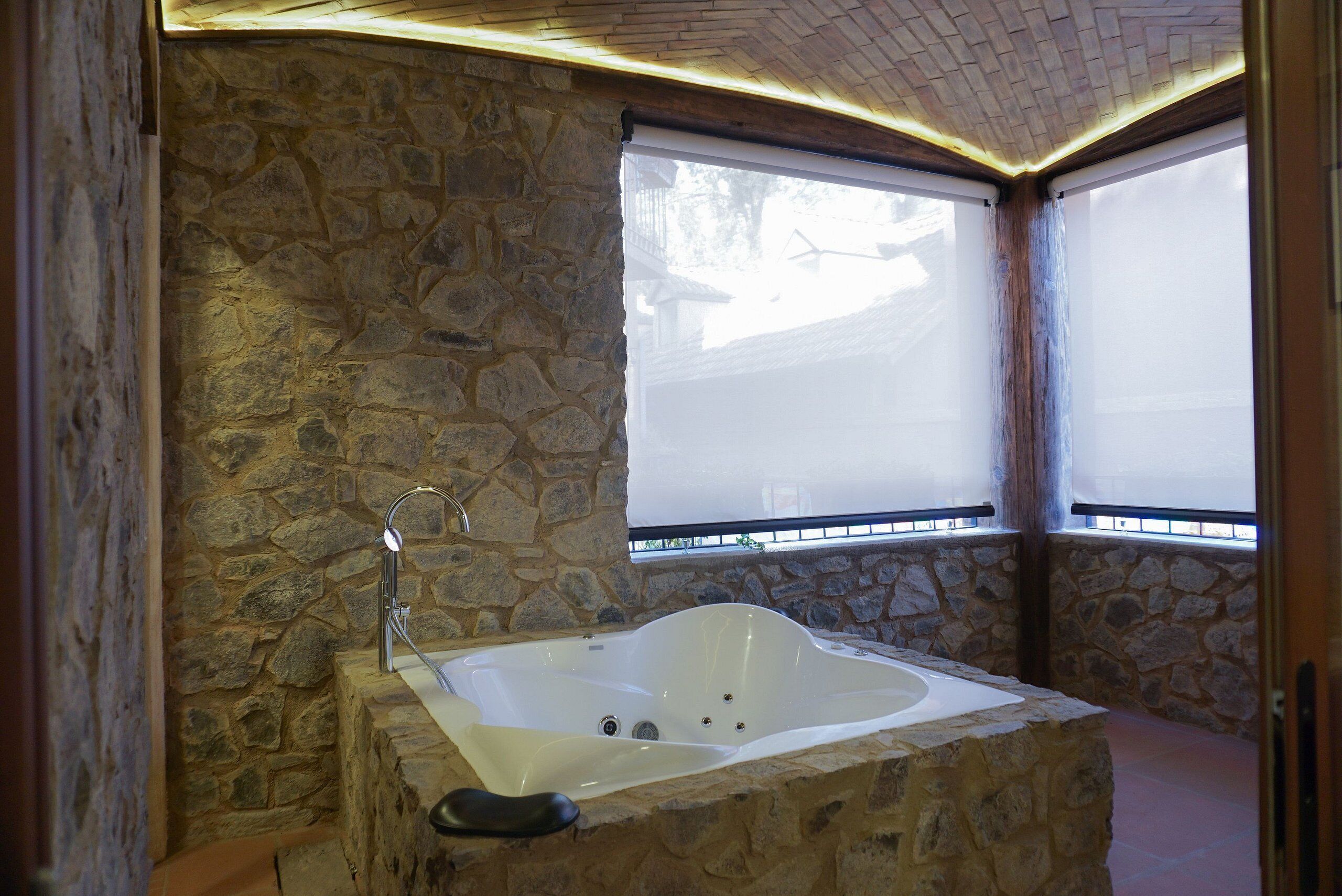 A stone-walled bathroom with a freestanding bathtub, a showerhead, and a window with blinds.