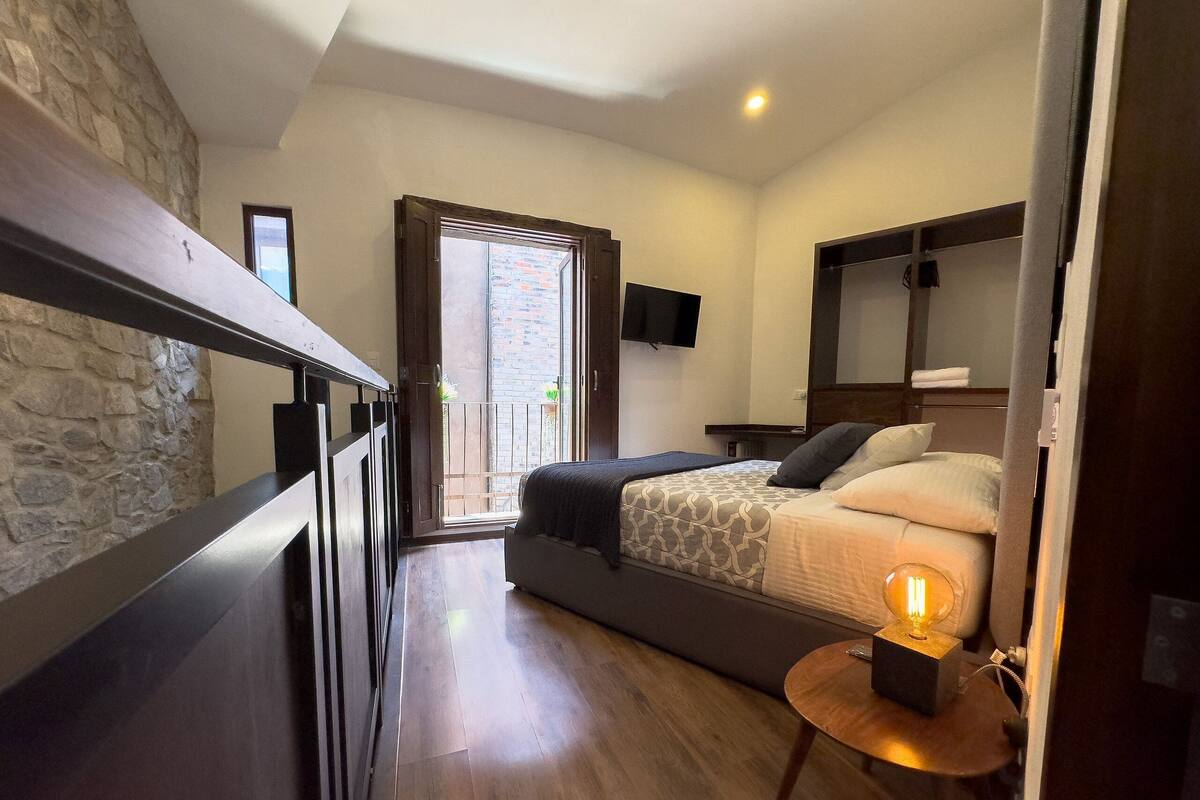 A bedroom with a bed, a small table with a lamp, a TV, and a balcony with a view of the city.