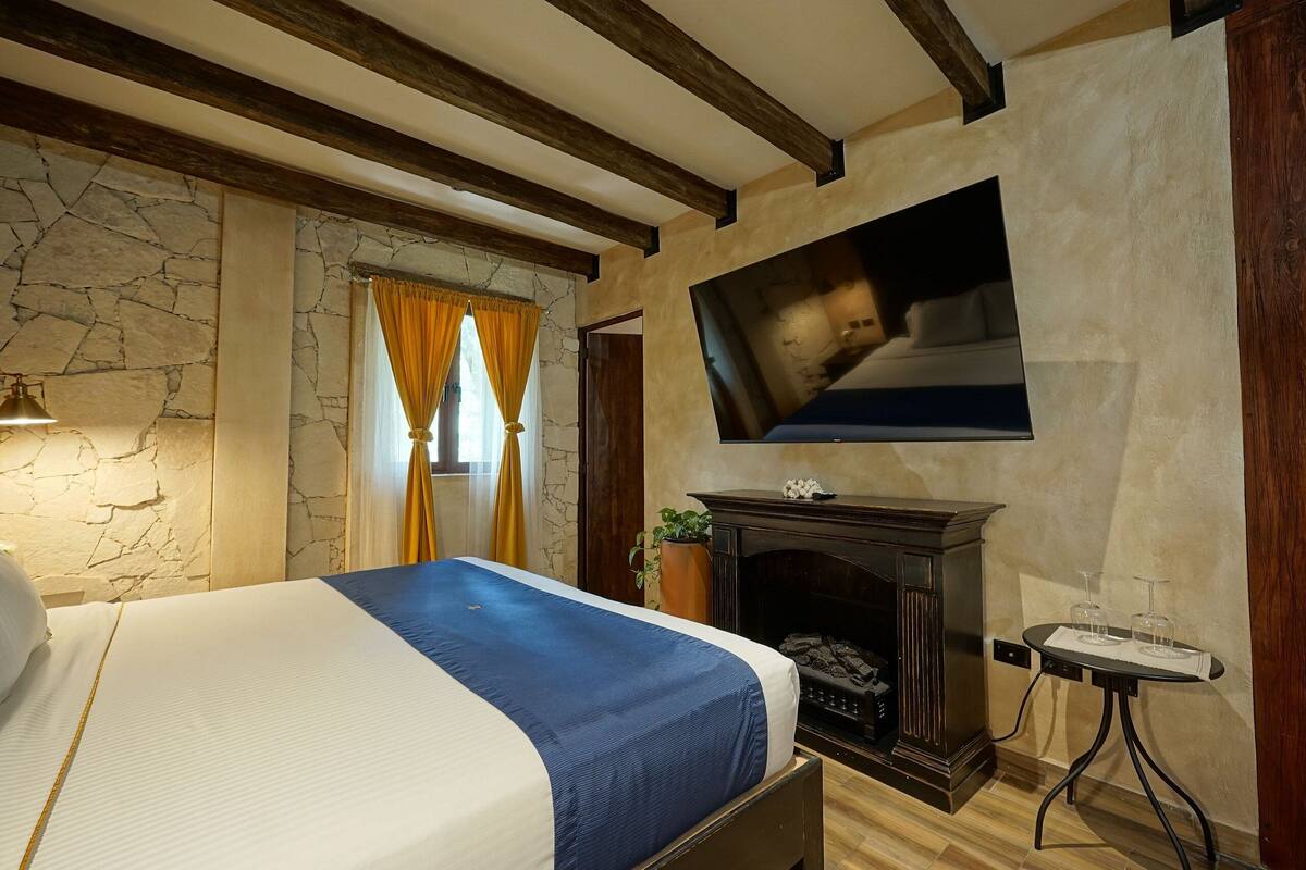 A bedroom with a bed, stone wall, wooden beams, a fireplace, a TV, and a small table with glasses.