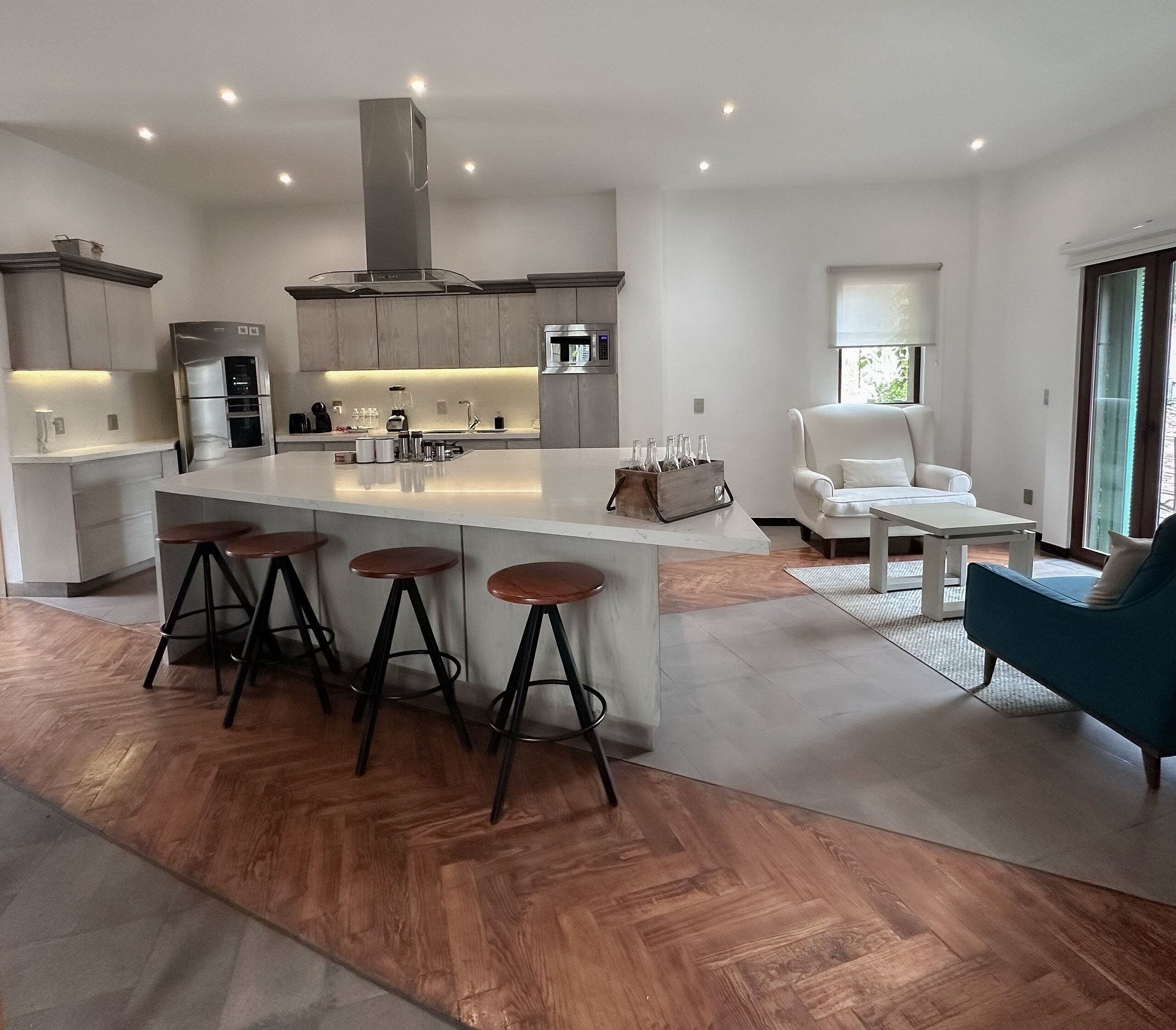 Een moderne keuken met een centraal eiland, barkrukken, een eethoek met een blauwe stoel en een groot raam.