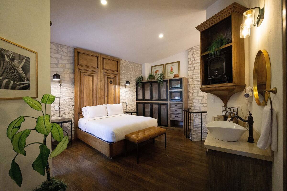 A bedroom with a bed, wooden wardrobe, stone wall, and a bathroom with a sink and mirror.