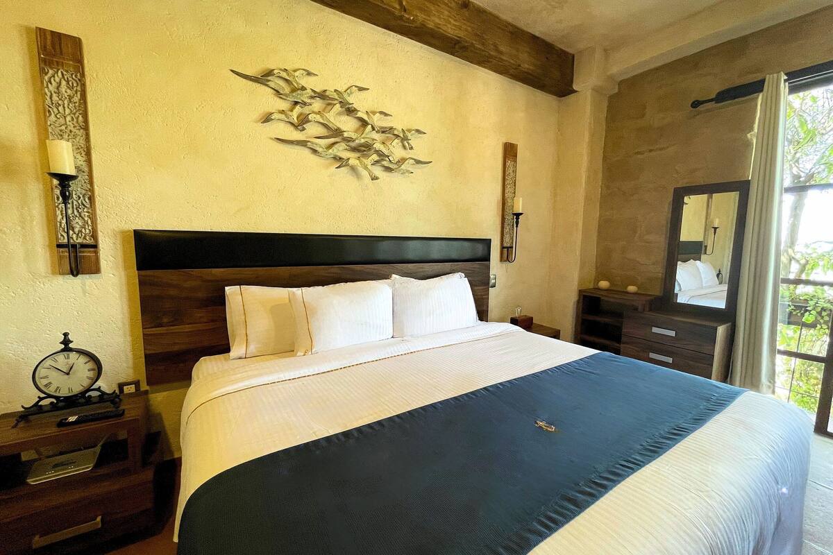 A neatly made bed with a dark blue bedspread, a wooden headboard, and white bedding. There is a wall clock on a bedside table, a decorative wall piece above the bed, and a mirror on the wall to the right.