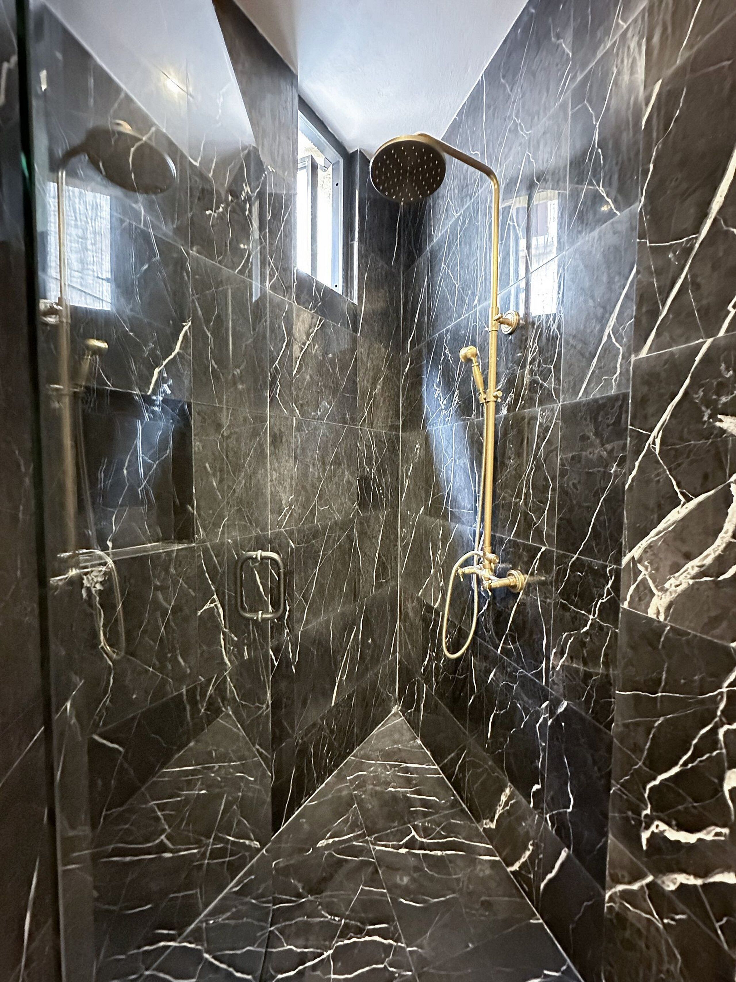 Une salle de bain moderne avec des murs et un sol en marbre foncé, une pomme de douche dorée et une fenêtre bien visible.