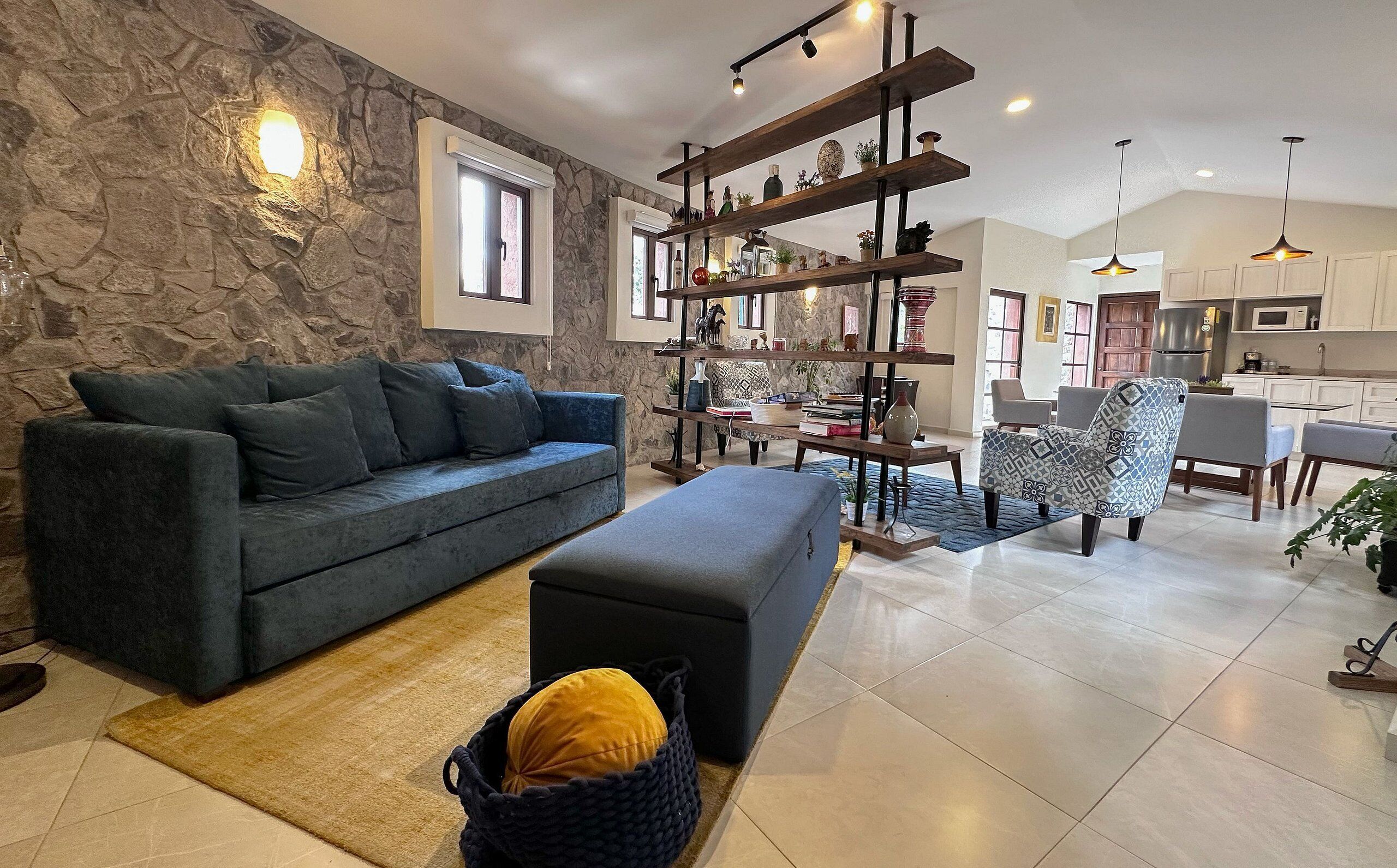 A modern living room with a stone wall, a dark blue sofa, a wooden coffee table, and a kitchen in the background.