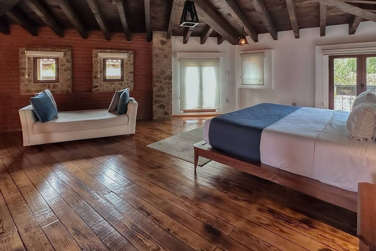 A rustic bedroom with exposed wooden beams, a stone wall, a wooden floor, a bed, a sofa, and two windows.