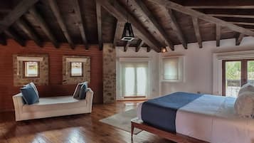 A rustic bedroom with exposed wooden beams, a stone wall, a wooden floor, a bed, a sofa, and two windows.