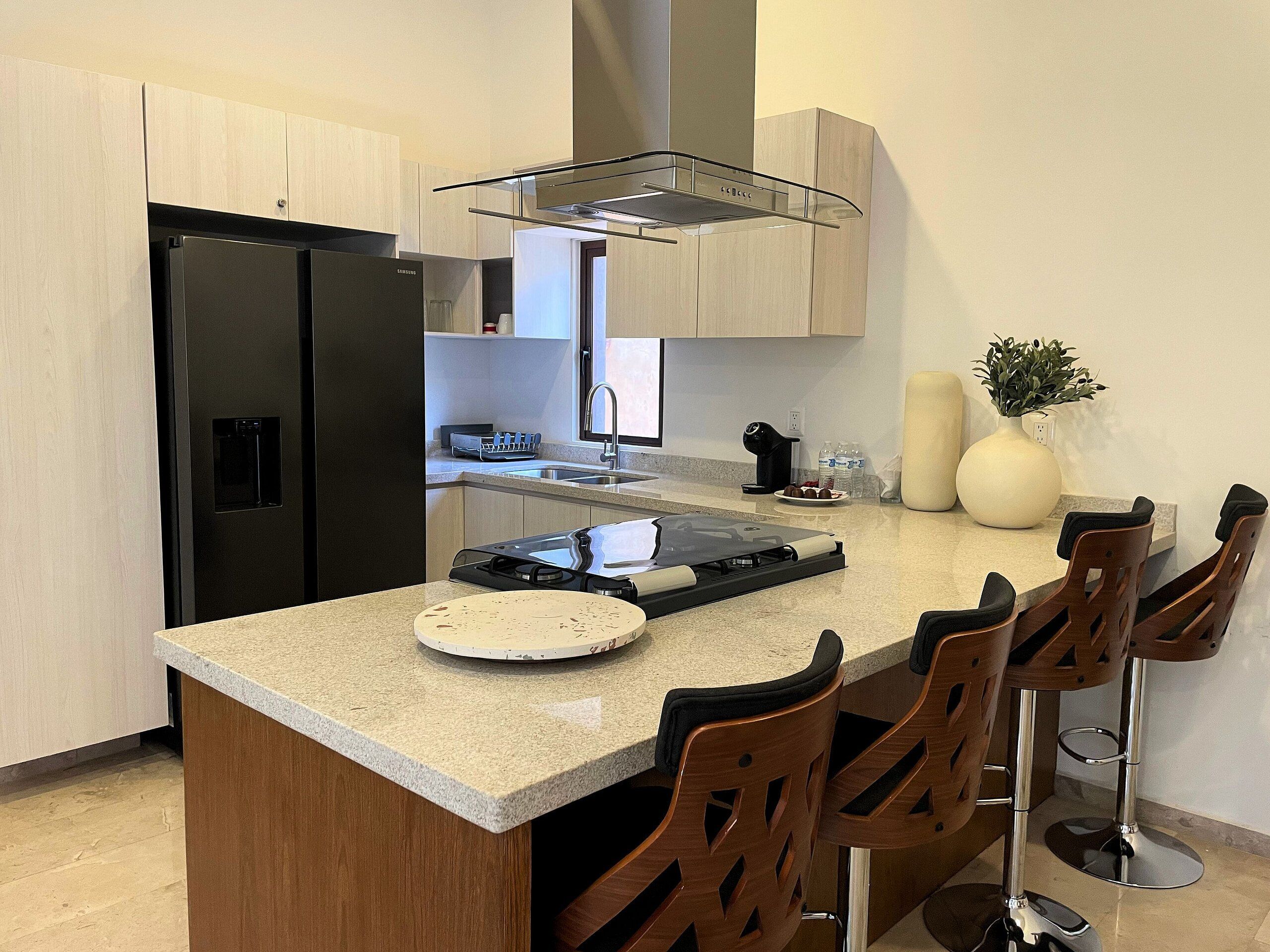 A modern kitchen with a built-in refrigerator, a stove, and a sink. There are bar stools and a vase with a plant on the counter.