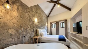 Un baño moderno con pared de piedra, una bañera independiente, una cama y un televisor sobre una estantería empotrada.