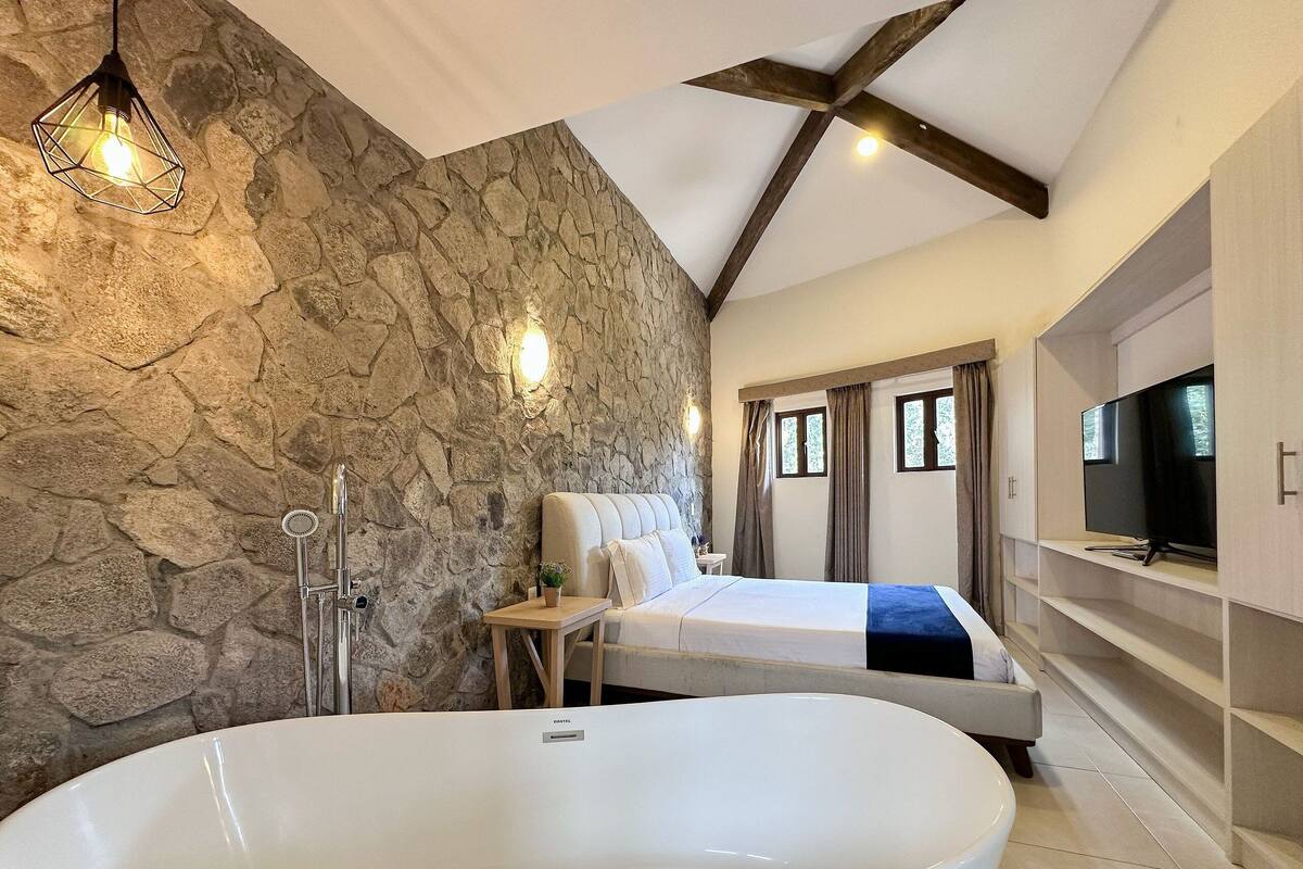 A modern bathroom with a stone wall, a freestanding bathtub, a bed, and a TV on a built-in shelf.