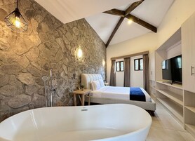 Un baño moderno con pared de piedra, una bañera independiente, una cama y un televisor sobre una estantería empotrada.