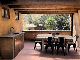 Un área de cocina al aire libre con una mesa y sillas de madera, un fregadero y vistas a los árboles y edificios circundantes.