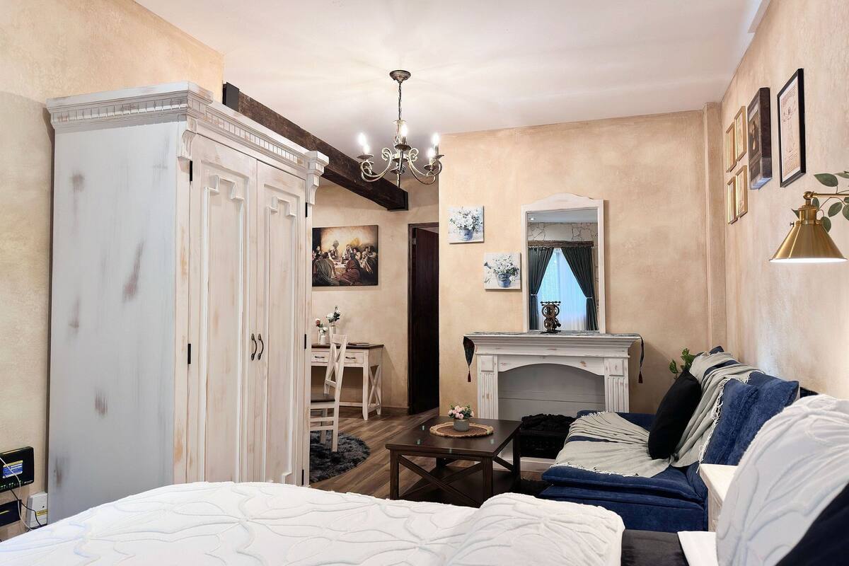 A bedroom with a large bed, a sofa, a dining table, and a fireplace.
