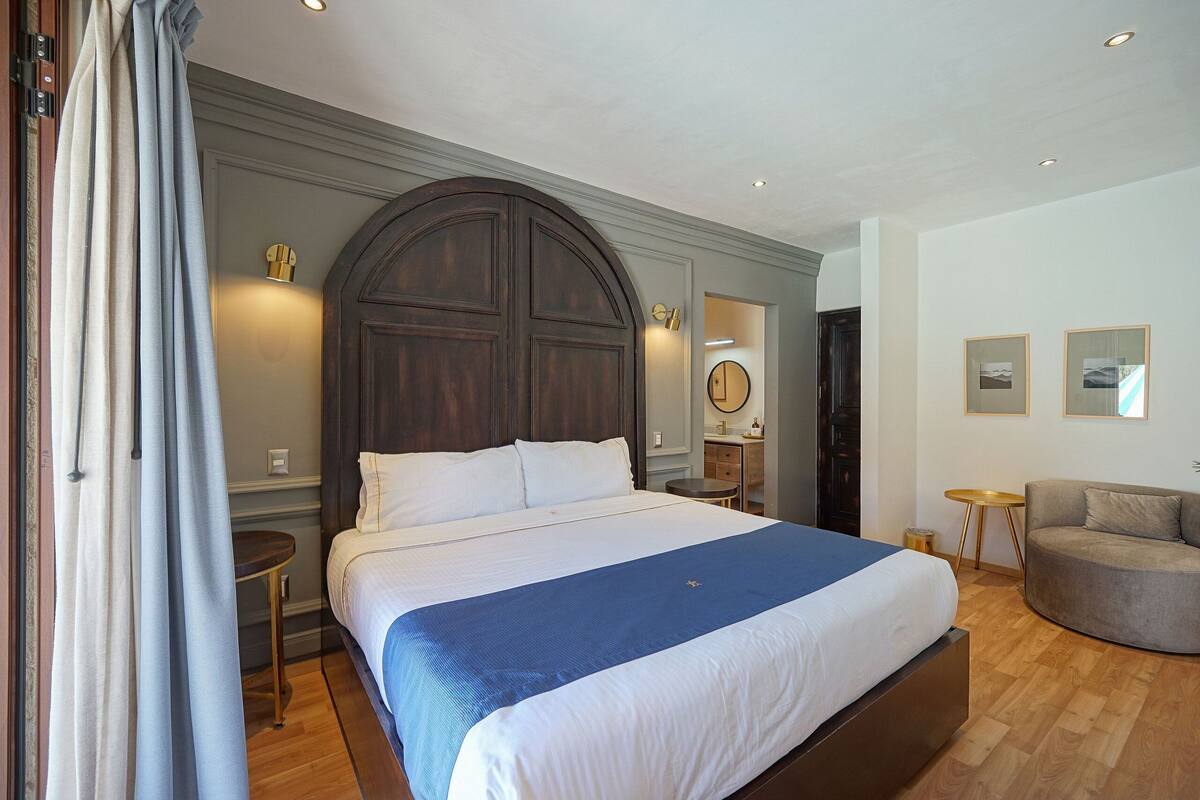 A large bed with a wooden headboard, a round mirror, a small round table, and a sofa in a well-lit room with wooden flooring.