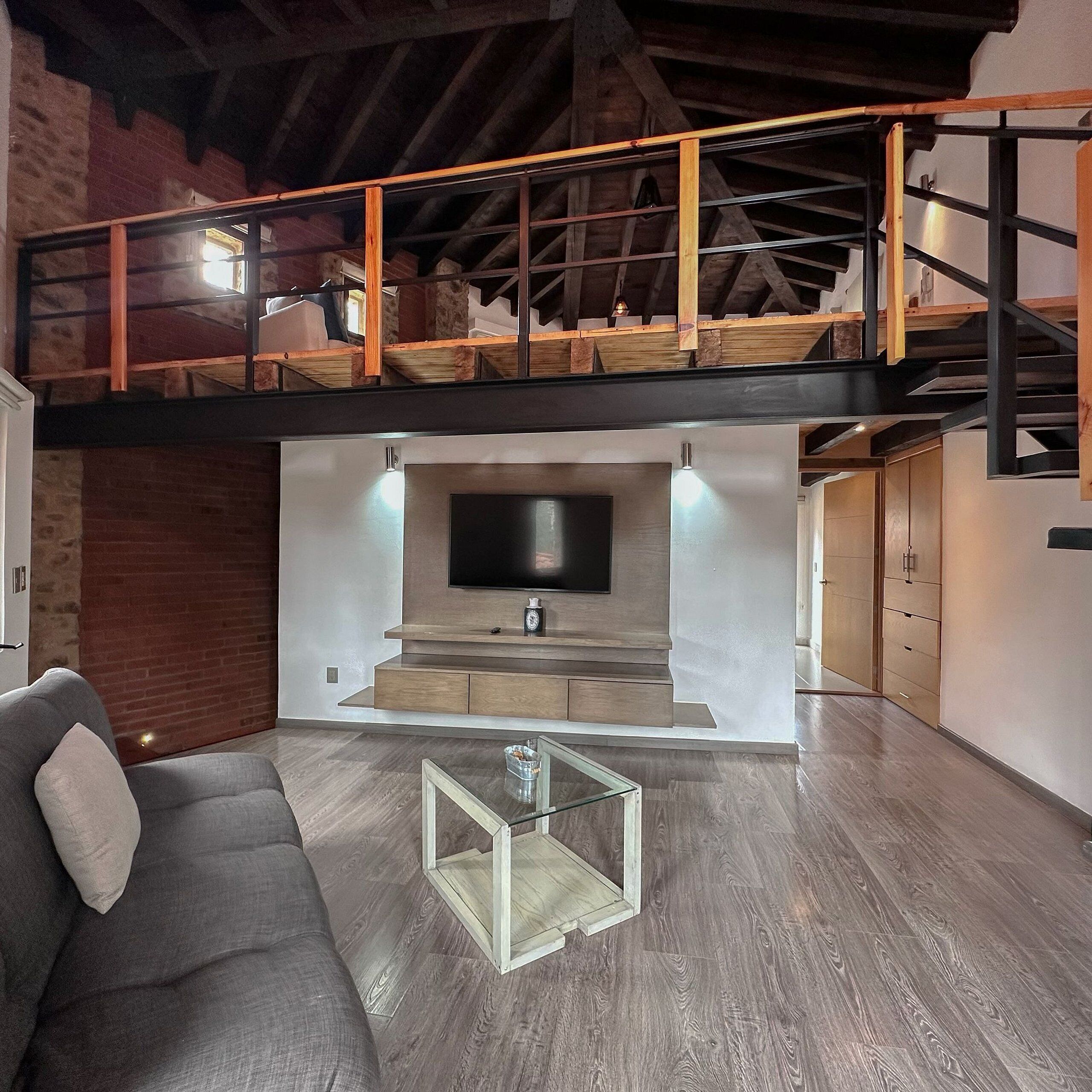 Un moderno espacio tipo loft con techo alto, vigas a la vista, televisor integrado y una escalera que conduce a un nivel superior.