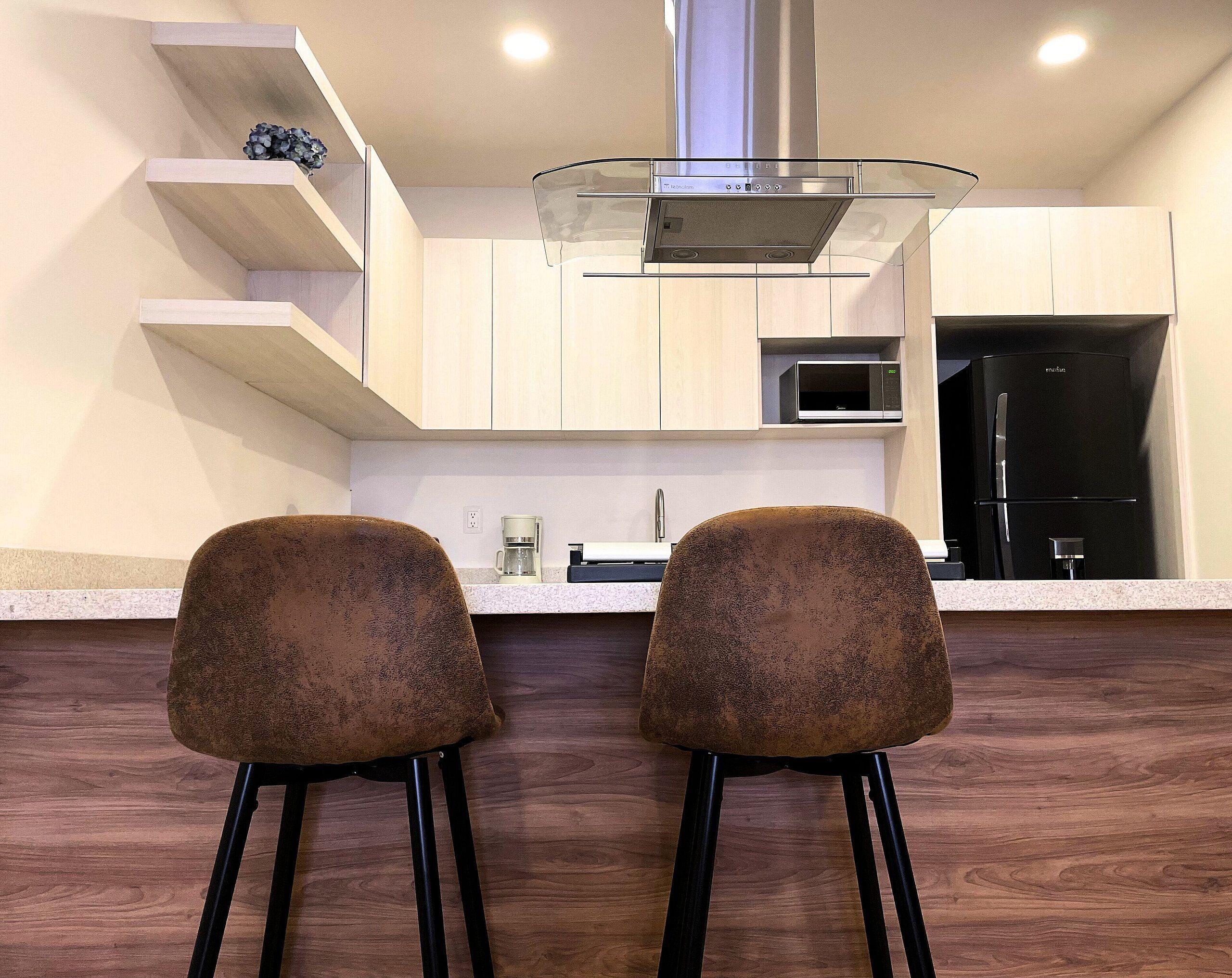 A modern kitchen with a stainless steel range hood, a microwave, and a black refrigerator. There are two bar stools with a textured seat and black legs.