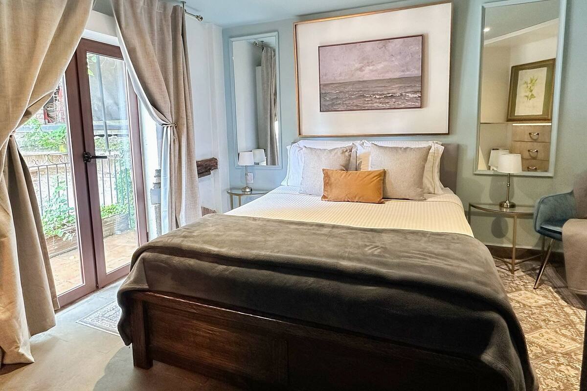 A bedroom with a large bed, a bedside table, a mirror, and a view of the outdoors.