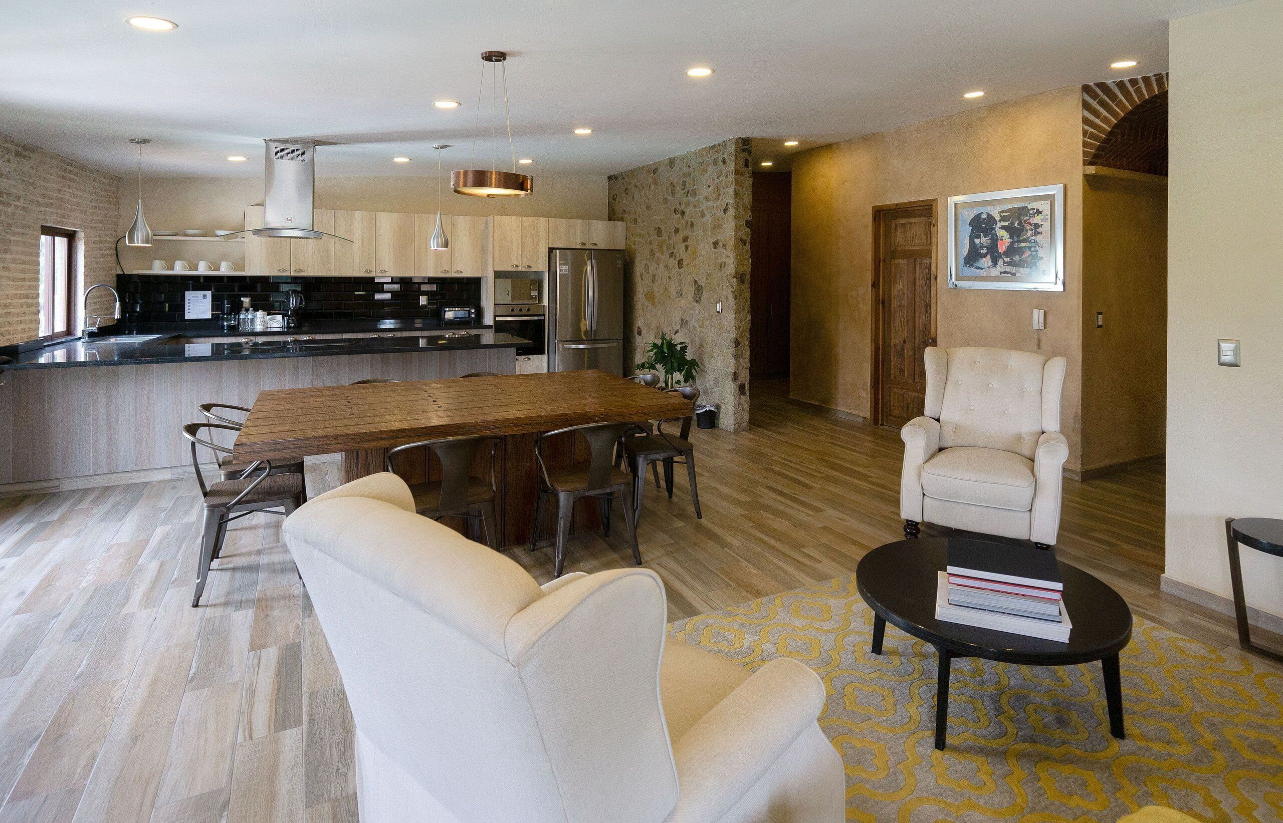 Una cocina moderna con una isla central, un comedor con mesa y sillas de madera, y una sala de estar con sofá y sillón.
