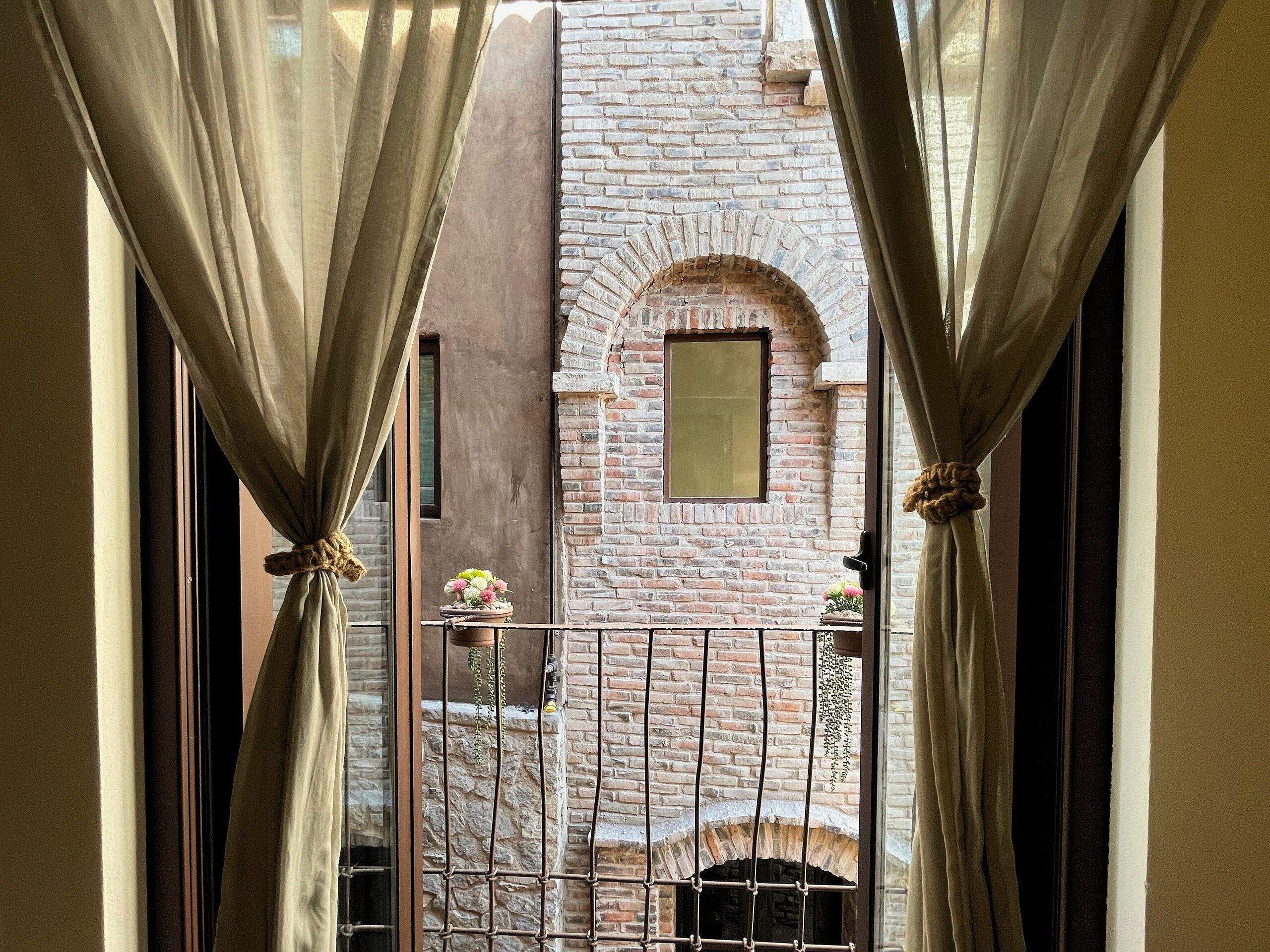 A view through a window with curtains, revealing a brick wall and a small window.