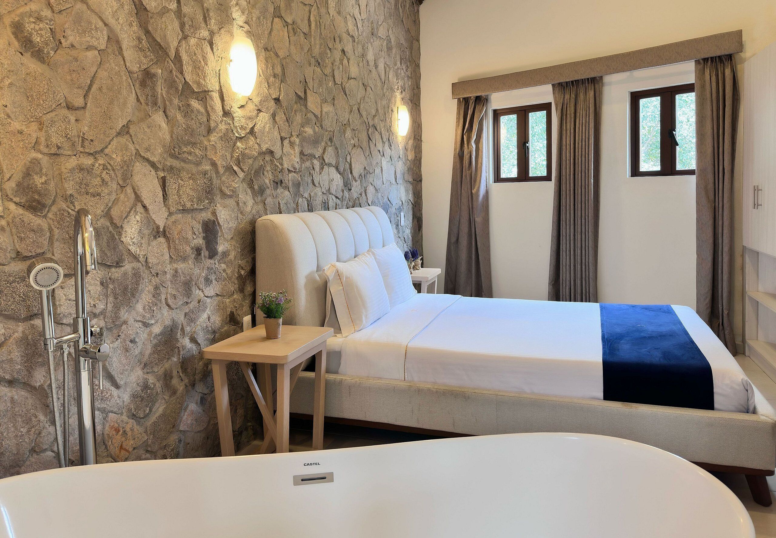A bedroom with a stone wall, a bed with a blue accent, a wooden nightstand, a white bathtub, and a window with curtains.