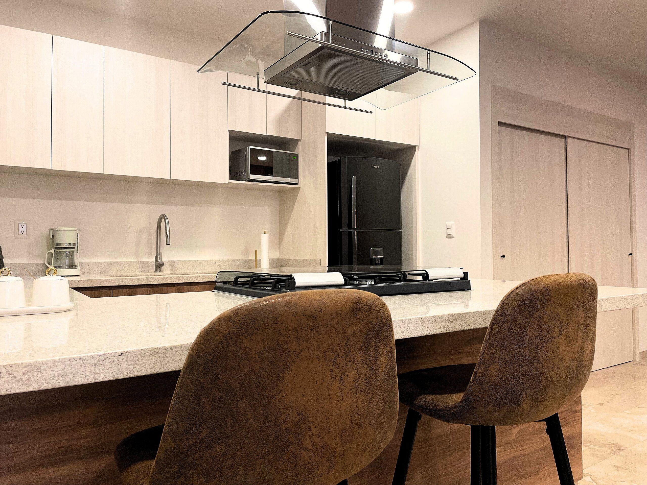 A modern kitchen with a stainless steel range hood, a microwave, and a black refrigerator. There are two chairs with a speckled pattern and a white countertop.