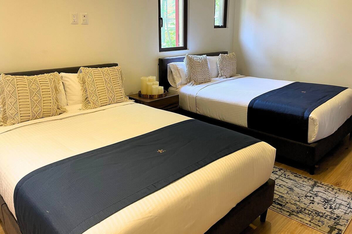 A double bed with a black headboard and a single bed with a dark headboard in a room with a small window and a nightstand with a lamp.