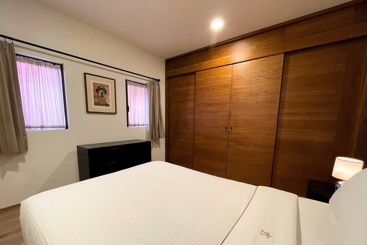 A bedroom with a bed, a wooden wardrobe, a TV, and two windows with curtains.