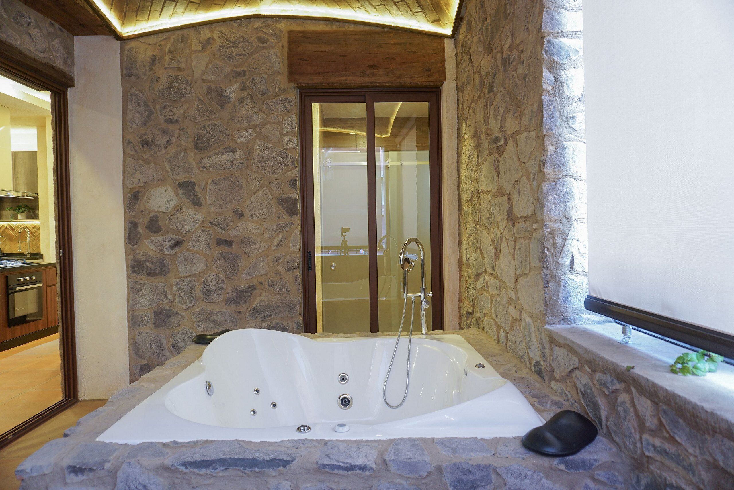 Un baño de paredes de piedra con una bañera grande, una ducha y techo de madera.