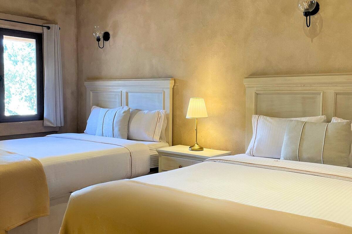 A hotel room with two beds, a nightstand with a lamp, and a window with curtains.