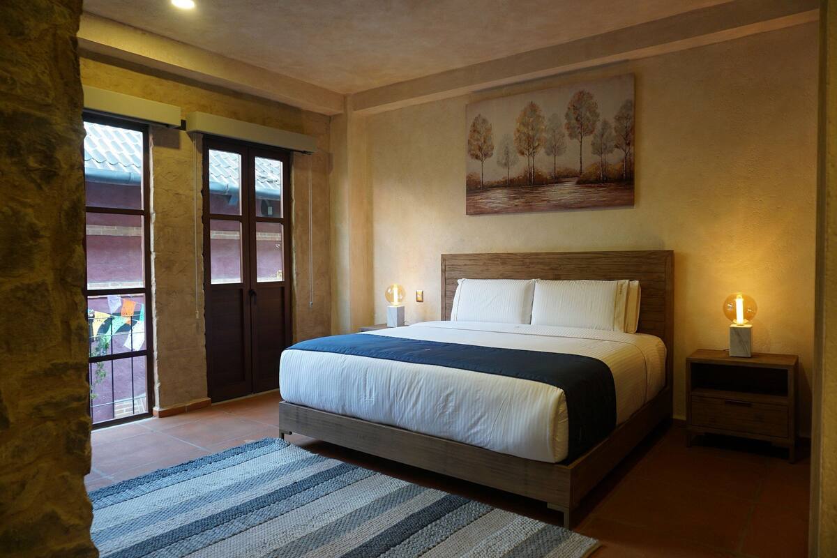 A bedroom with a large bed, a painting on the wall, and a nightstand with a lamp.