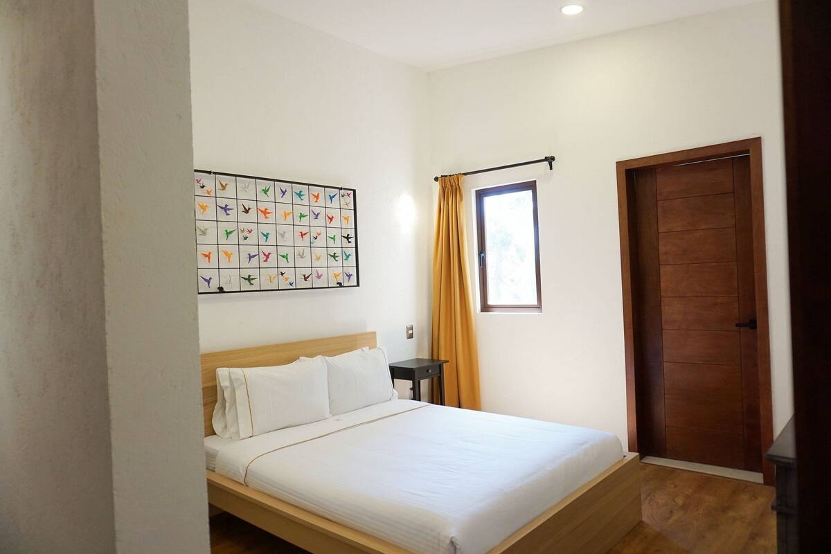 A bedroom with a bed, a nightstand, a window with curtains, a door, and a wall art piece.