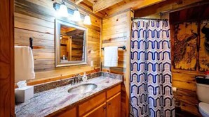 Une salle de bain rustique, avec une cabine en bois, un lavabo avec un plan de travail en granit, un miroir et un rideau de douche à motifs.