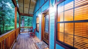 Une terrasse en bois avec un paillasson, une porte ornée d’un panneau de verre décoratif et une vue sur la forêt environnante.