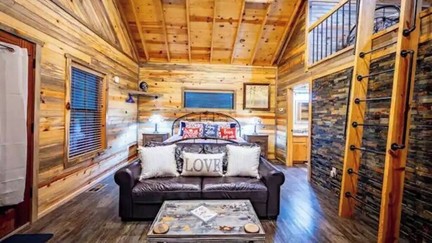Un intérieur de chalet chaleureux avec un plafond en bois, un canapé en cuir agrémenté de coussins ornés du mot « AMOUR », une table basse et un mur en pierre.