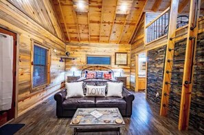 A cozy cabin interior with a wooden ceiling, a leather sofa with 'LOVE' pillows, a coffee table, and a stone wall. - Travelin Lights;Perfect Cabin Experience- Sleeps 4 (Broken Bow)