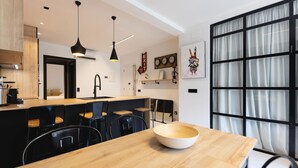 A modern kitchen with a wooden countertop, black chairs, and pendant lights. There is a large window with curtains and a view of the outside.