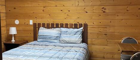 A bedroom with a wooden headboard, a bed with striped bedding, a nightstand with a lamp, and a chair.