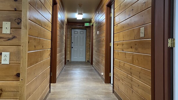 Un pasillo de madera con una puerta al final, una señal de salida y una lámpara en el techo.