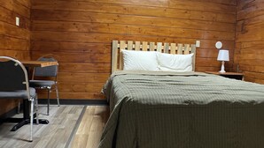 Una habitación con paredes de madera, una cama con colcha verde a rayas, una mesita de noche con lámpara y una silla.