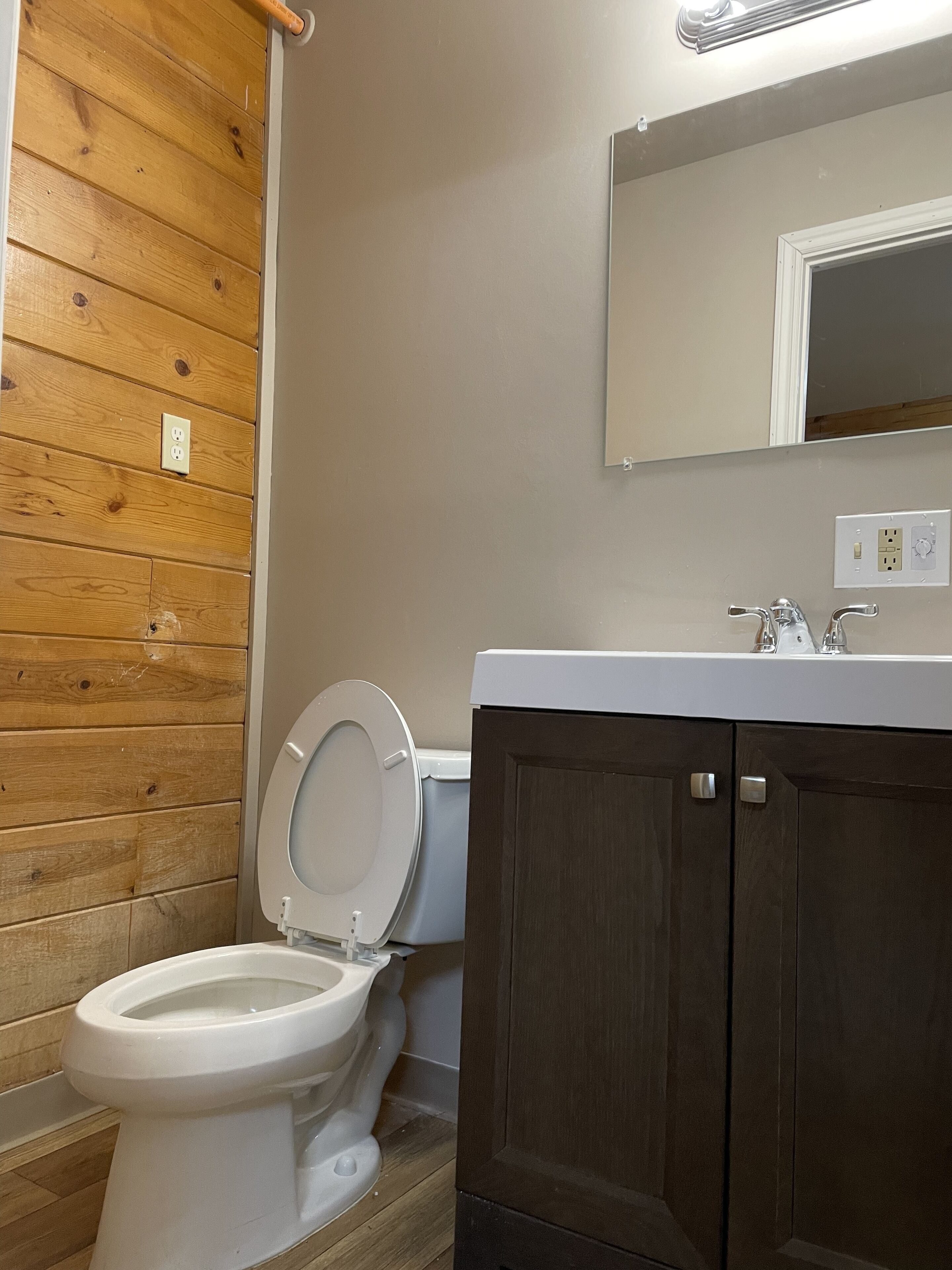 Een badkamer met een houten wand, een wit toilet, een donker houten kastje en een spiegel boven de wastafel.