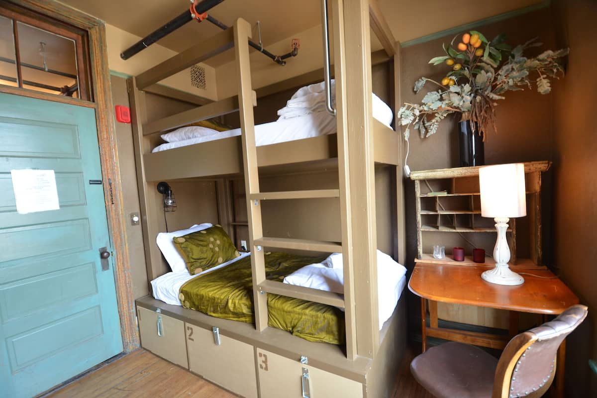 A bunk bed room with a desk and chair, a small table with a lamp, and a vase with flowers.