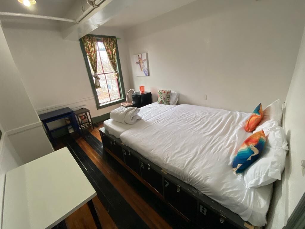 A compact hotel room with a single bed, a small table, a window with curtains, and a wall-mounted artwork.